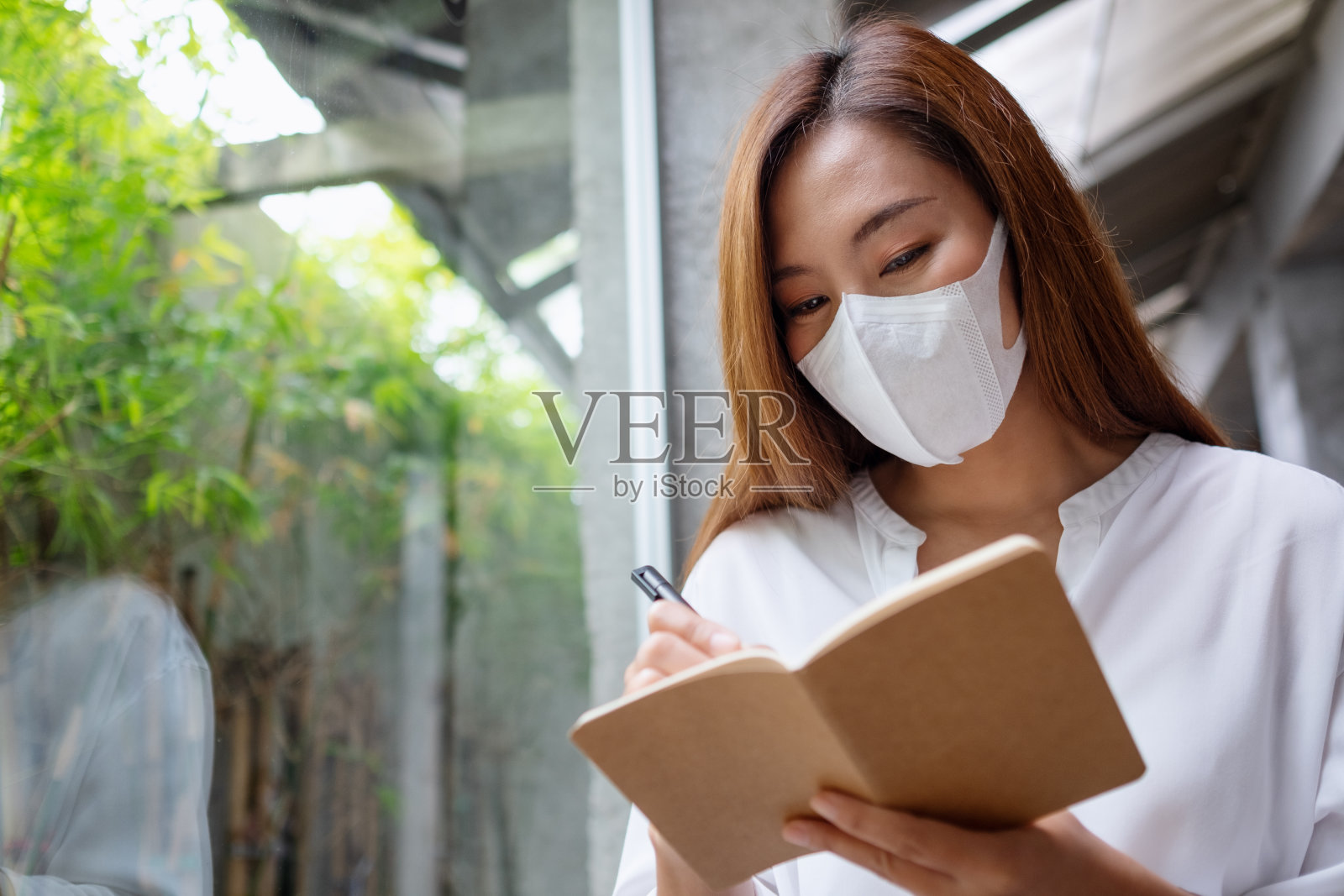一个年轻的亚洲女人带着防护面罩，拿着笔记本在上面写字照片摄影图片