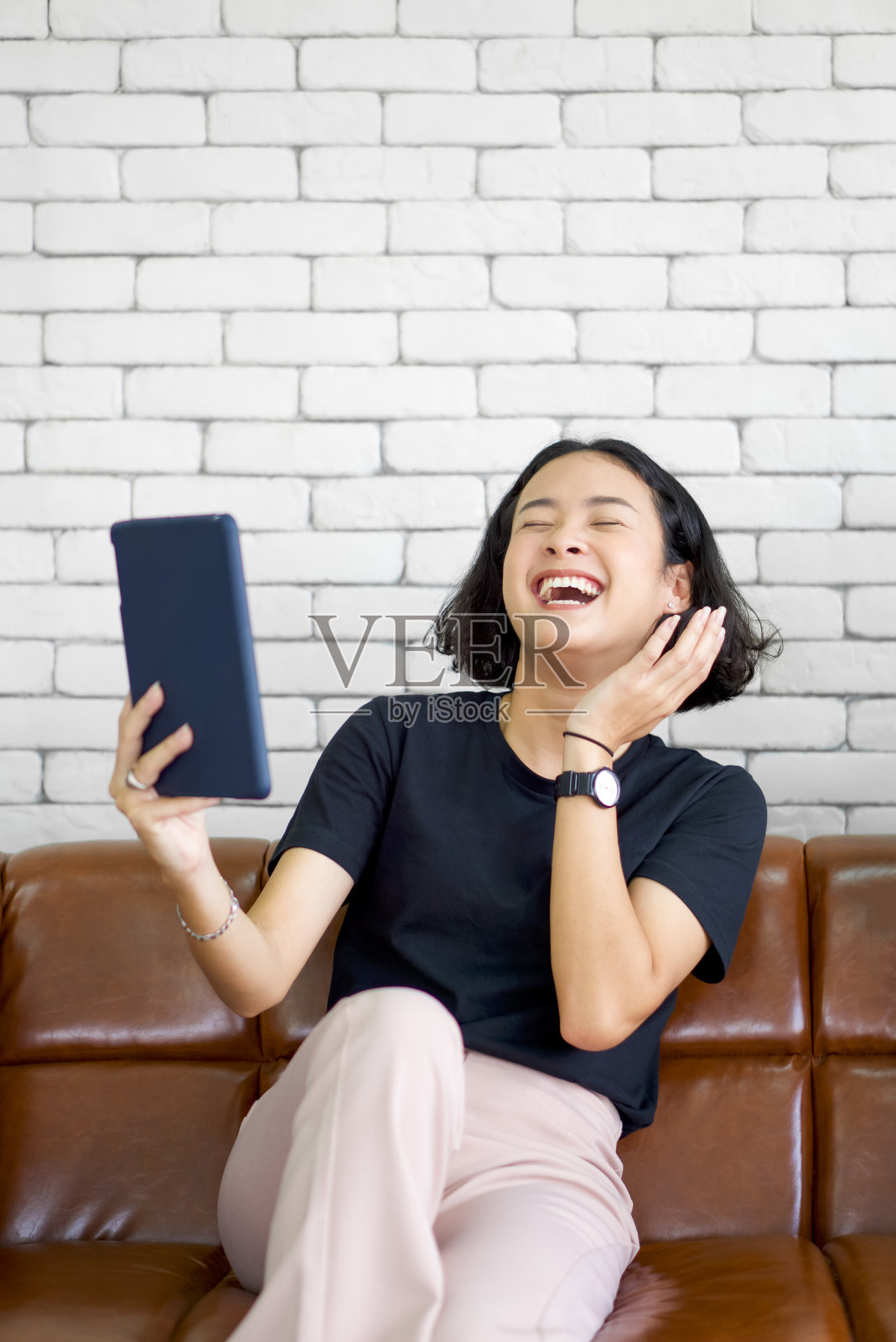 一名身穿黑色t恤的亚洲年轻女子坐在客厅沙发上，用平板电脑看喜剧片，笑得很开心。在家享受休闲周末。无压力的概念。照片摄影图片