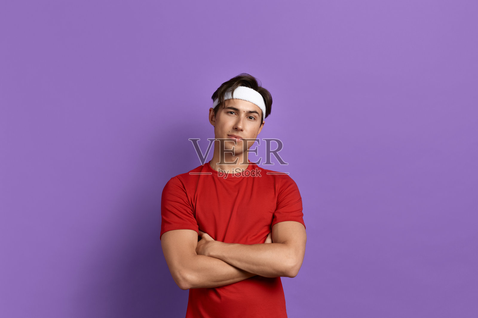 友好的年轻运动员或教练双臂交叉在胸前，是健身成瘾，提倡健康的生活方式，正在去工作，穿着红色的t恤和白色的头带，孤立在紫色的背景。照片摄影图片