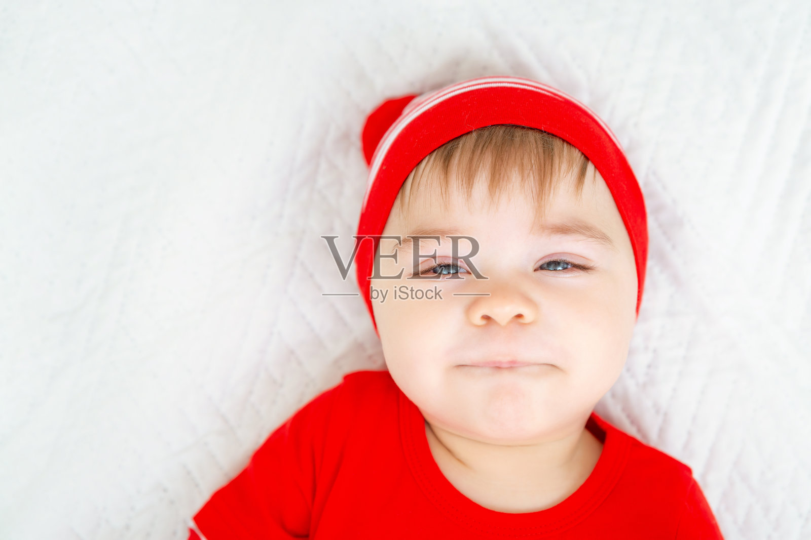 穿着圣诞衣服的小婴儿眯着眼睛傻笑。一个在白色背景上带着情绪的孩子的特写。看相机。冬天的假期。照片摄影图片