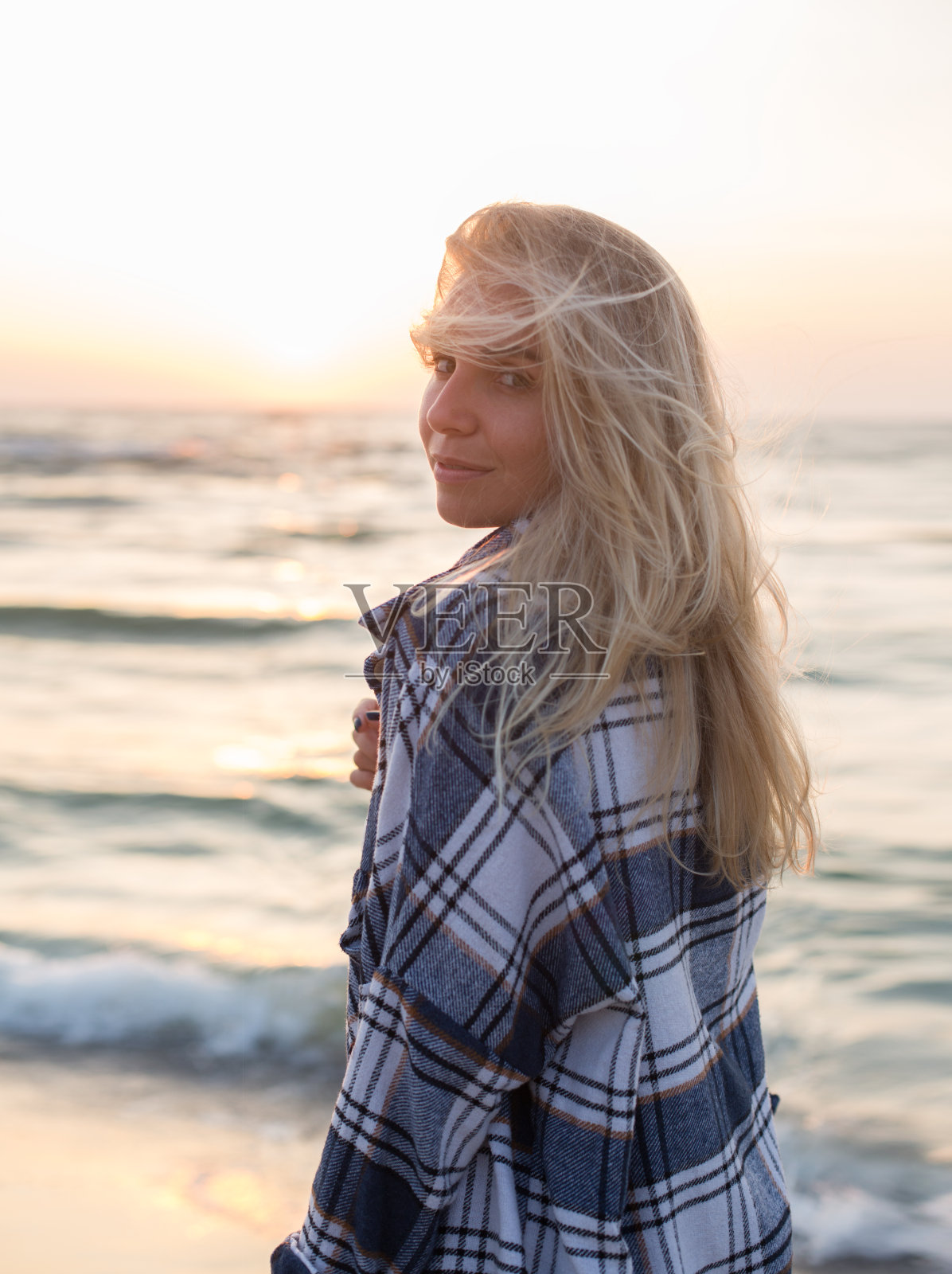 美丽快乐的金发女孩穿着运动衫赤脚站在日出的海滩上照片摄影图片