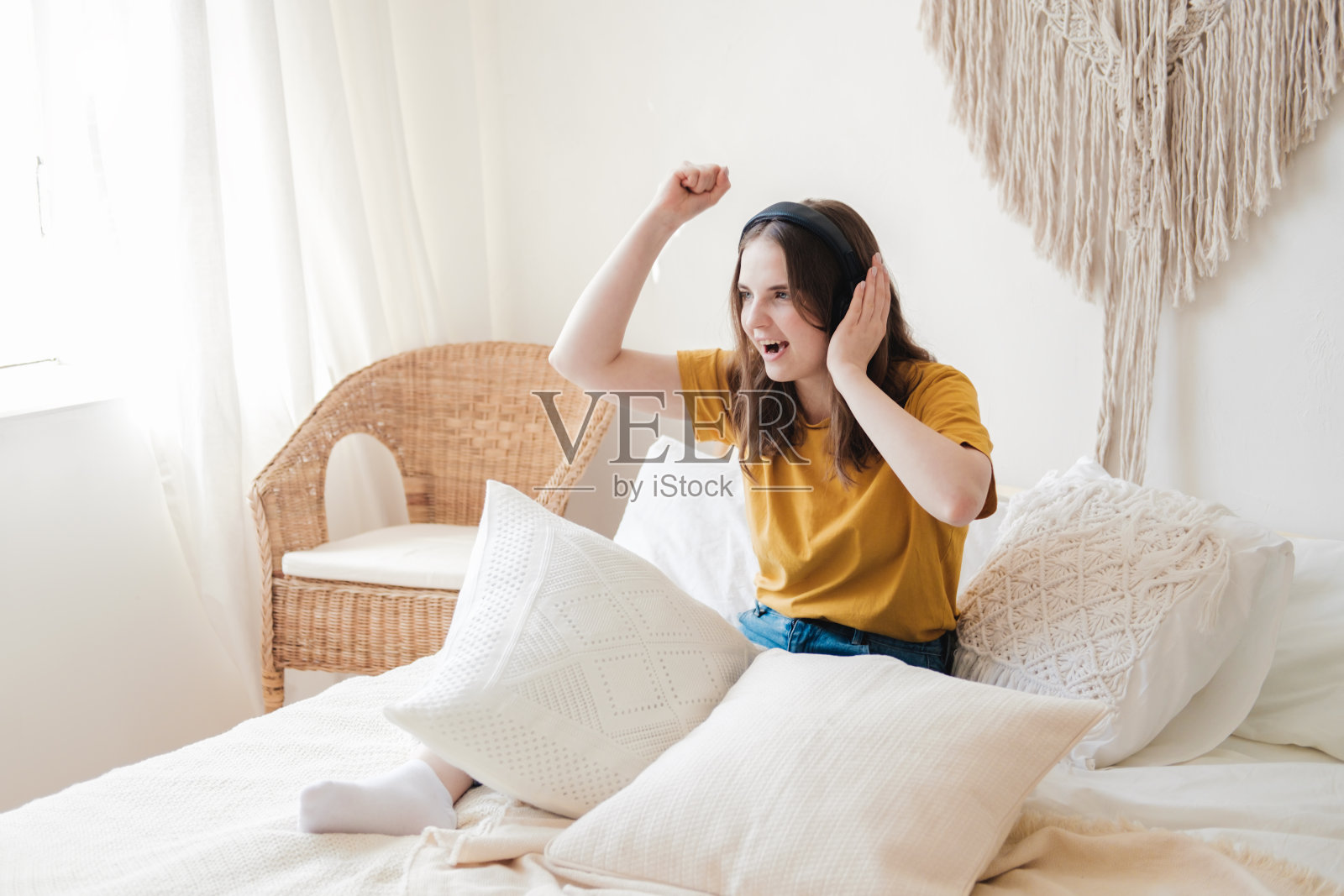 年轻漂亮的女学生穿着橙色t恤和蓝色牛仔裤，枕着枕头坐在床上，戴着耳机听有声书、音乐和播客。隔离期间在家自学的概念。照片摄影图片