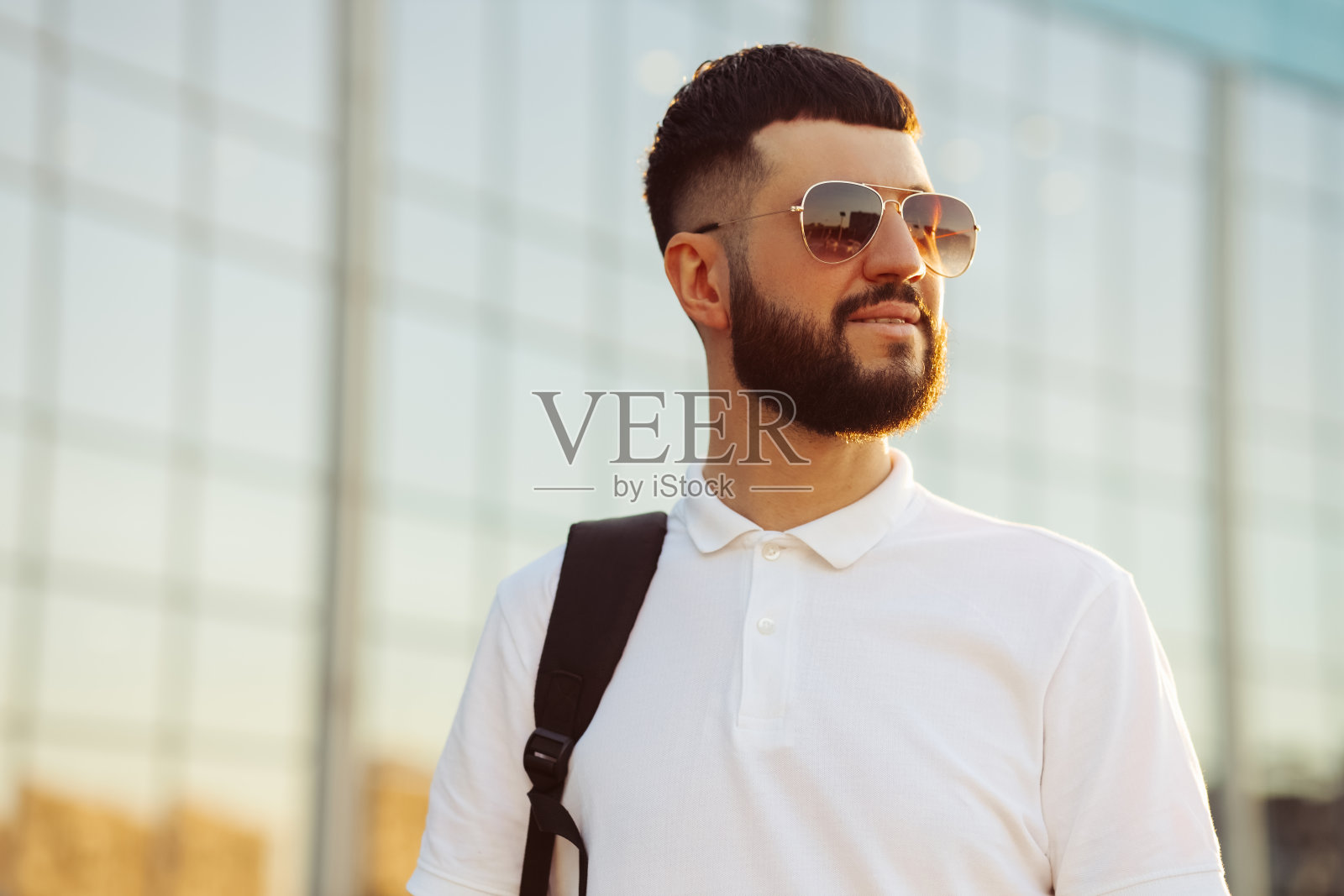 在一个阳光明媚的日子里，一名戴着太阳镜，留着胡子的男子，穿着一件休闲的白衬衫，微笑着。一个在室外玻璃建筑附近的人照片摄影图片