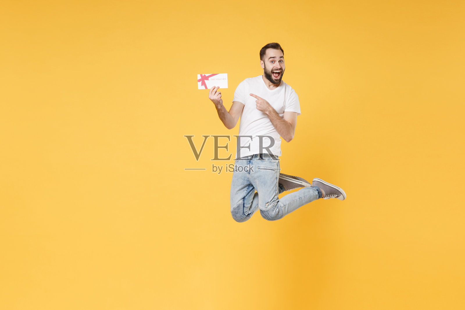 兴奋的年轻大胡子男人，男人在白色休闲t恤摆孤立的黄色墙壁背景工作室。人们生活方式的概念。模拟复制空间。跳着指着礼券上的食指。照片摄影图片