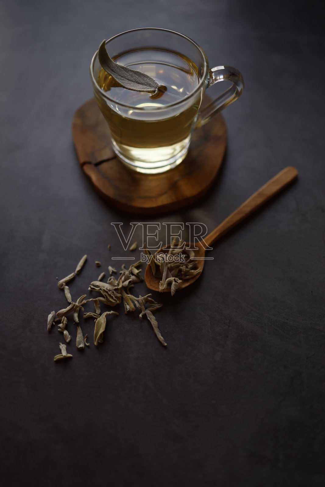 鼠尾草茶在玻璃杯与柄木制杯垫在黑色混凝土背景。有一片新鲜的鼠尾草茶叶放在鼠尾草茶上。杯子旁边是一个木勺和撒在地上的干鼠尾草茶。花草茶。照片摄影图片