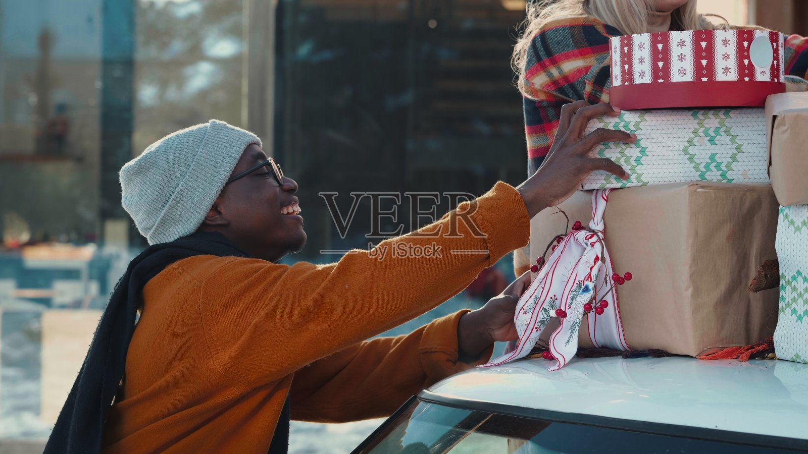 快乐的年轻非洲男人和白人女人在雪天的慢动作外面把礼物盒子放在车顶上。照片摄影图片