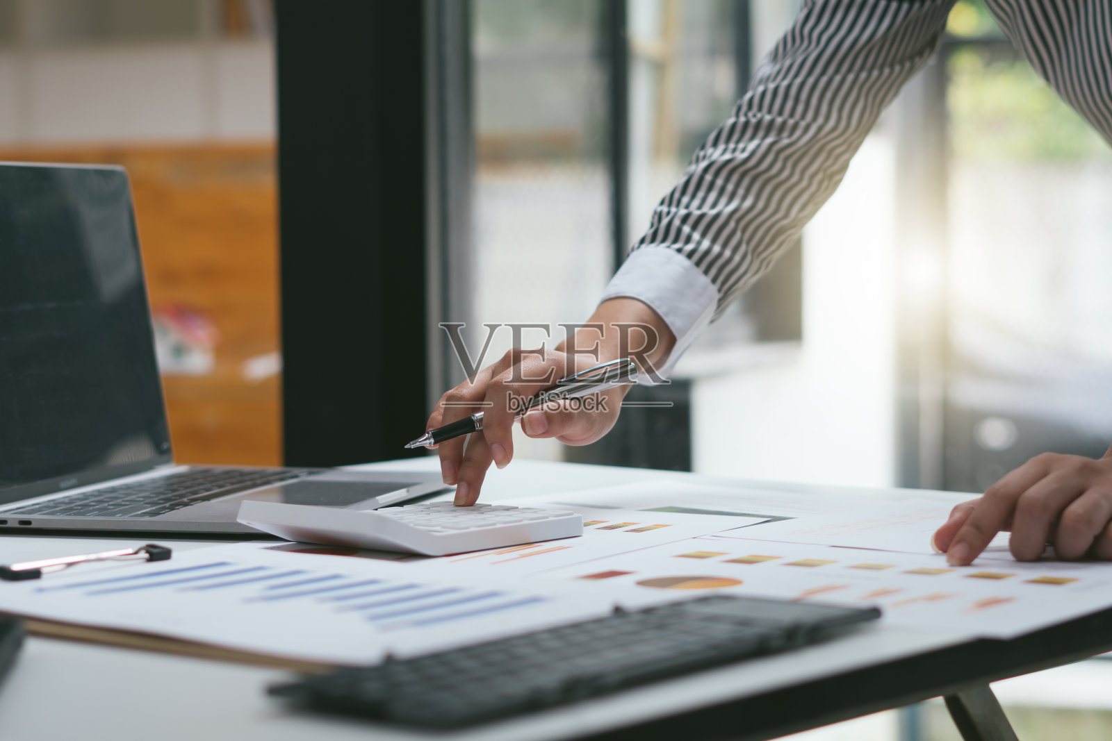 商人拿着笔和指示纸图表总结分析年度业务报告与使用笔记本电脑在房间办公桌。照片摄影图片