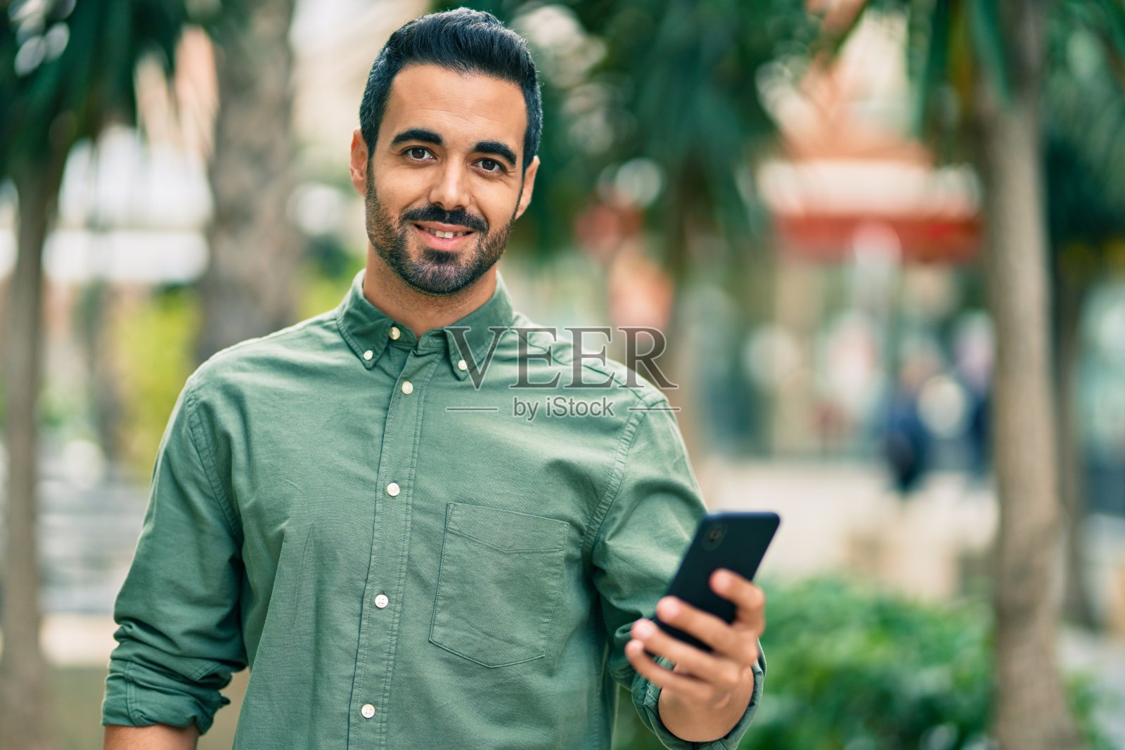 年轻的西班牙裔男子开心地在城市里使用智能手机。照片摄影图片