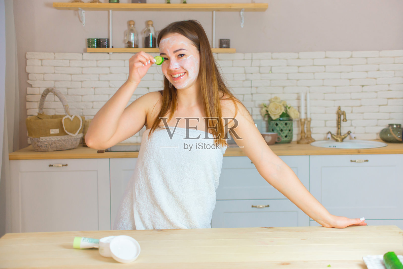一位妇女带着面膜和黄瓜片在家里的厨房里。面部和身体护理照片摄影图片
