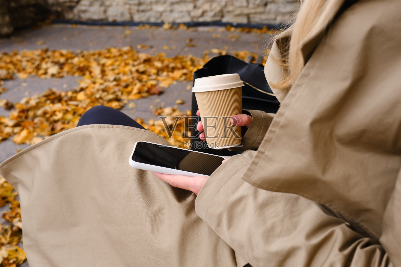 拿着智能手机和一杯咖啡的女孩坐在秋天公园的长椅上。一位女士正在发送短信或使用手机上的应用程序。在舒适的散步中，用纸杯热饮照片摄影图片