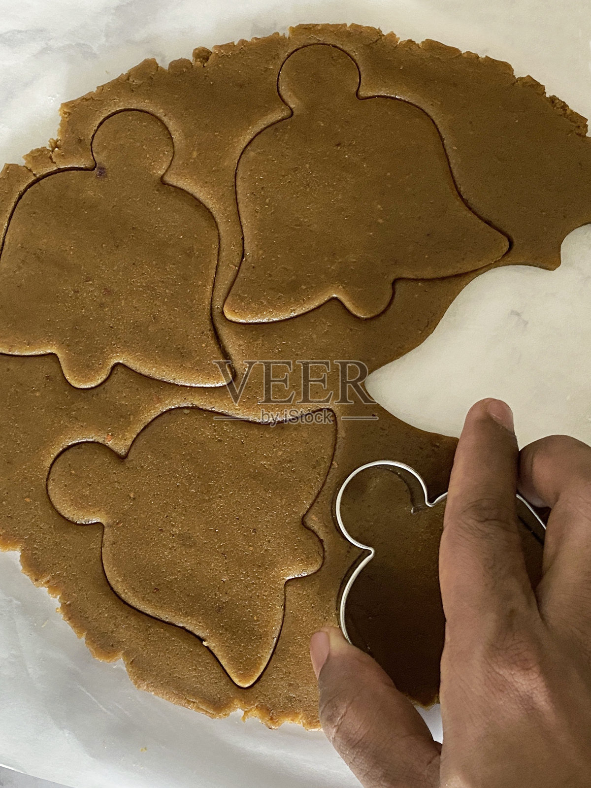 特写画面:一个不认识的人正在用金属钟饼干刀切自制的饼干，姜饼曲奇面团滚在防油的羊皮纸上，圣诞钟被切出形状，家庭烘焙，高架视野照片摄影图片