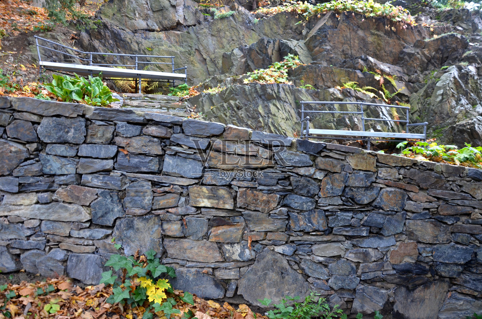 不规则几何多边形的木凳放在公园的石灰色路面上，低矮的挡土墙堆叠着斑岩。带有楼梯和自然岩石的观赏性花园照片摄影图片