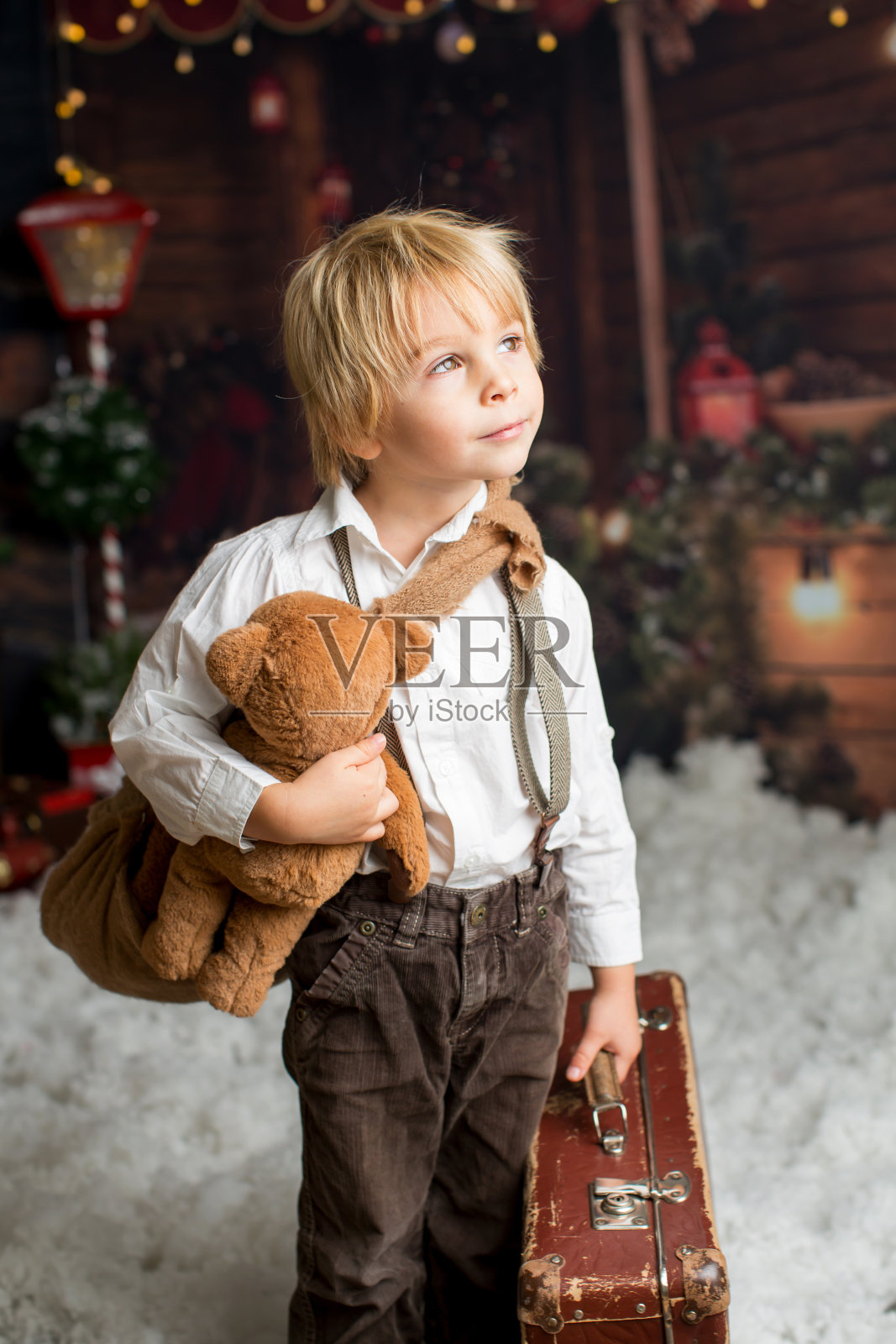 可爱时尚的小男孩，在雪地里和泰迪熊在木屋的圆木前玩耍照片摄影图片