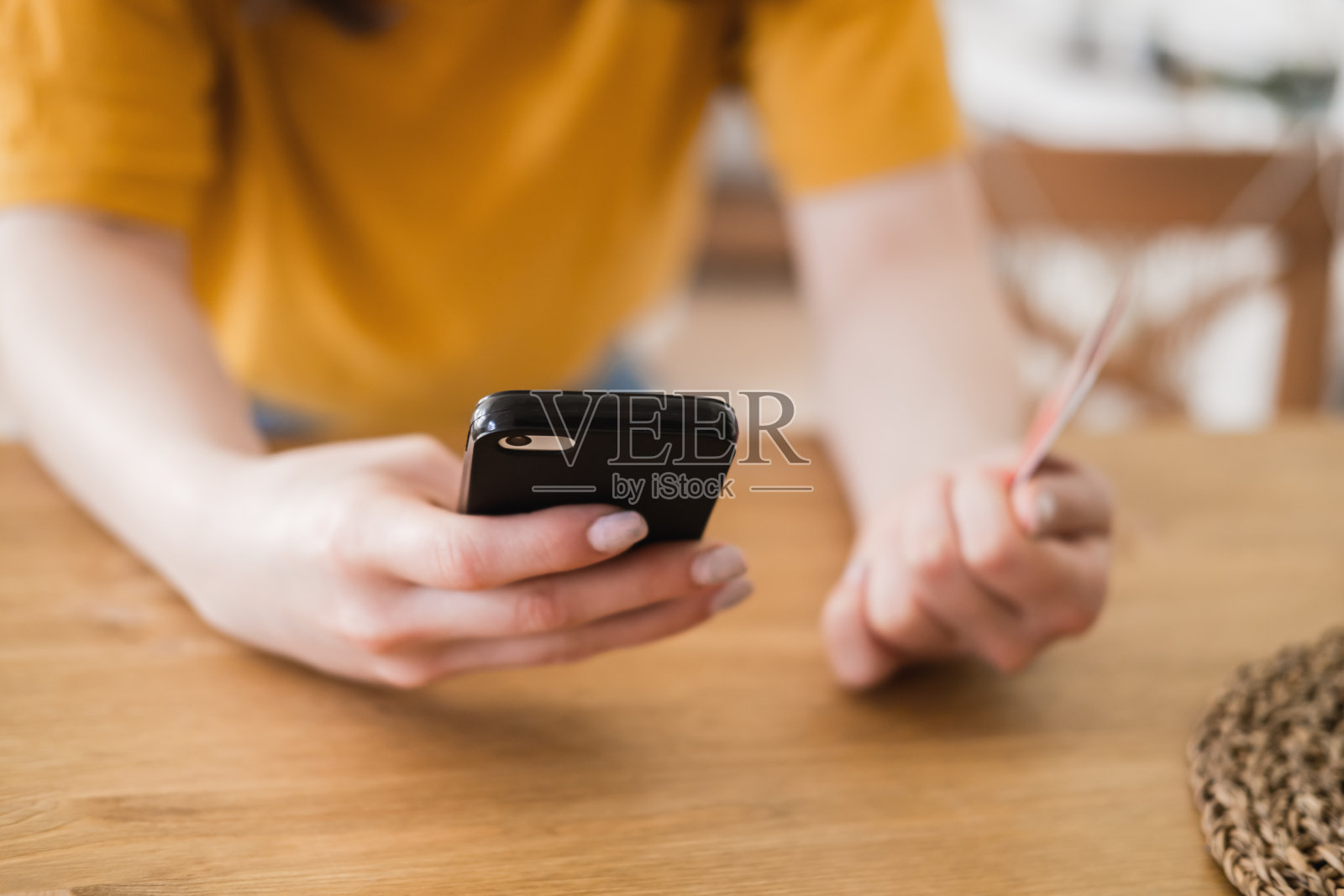 穿着亮黄色t恤的可爱年轻女子坐在桌旁，用智能手机和信用卡在网上购物。冠状病毒大流行期间的网上购物。照片摄影图片