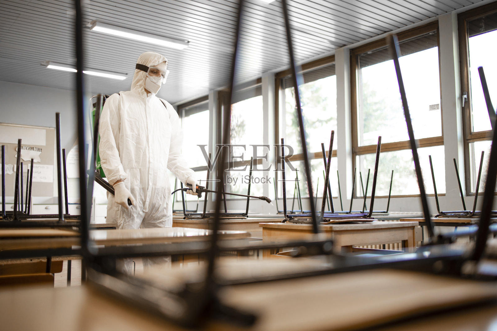 冠状病毒大流行期间，穿着白色无菌防护服的工作人员在学校教室对桌椅进行消毒消毒。照片摄影图片