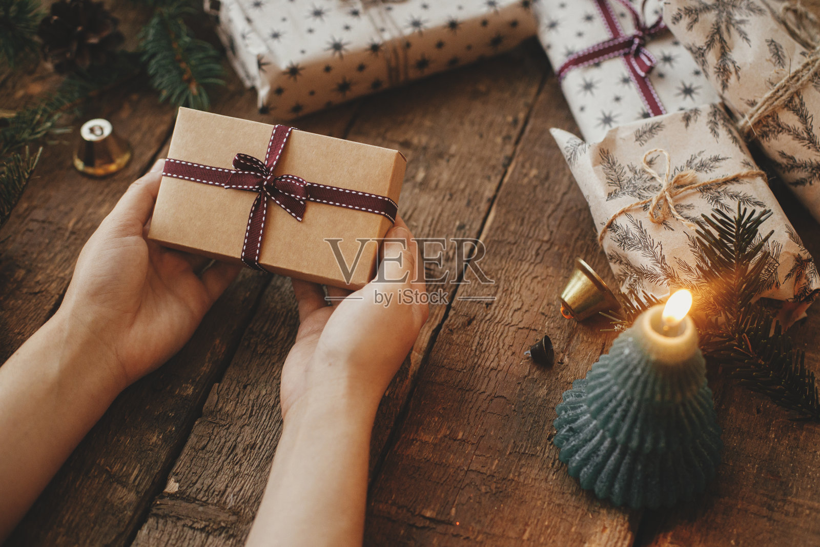 圣诞礼物。质朴的木桌上摆着蜡烛、剪刀和冷杉树枝，双手捧着时髦的圣诞礼物。时尚的斯堪的纳维亚圣诞礼物，大气的穆迪形象。圣诞快乐!照片摄影图片