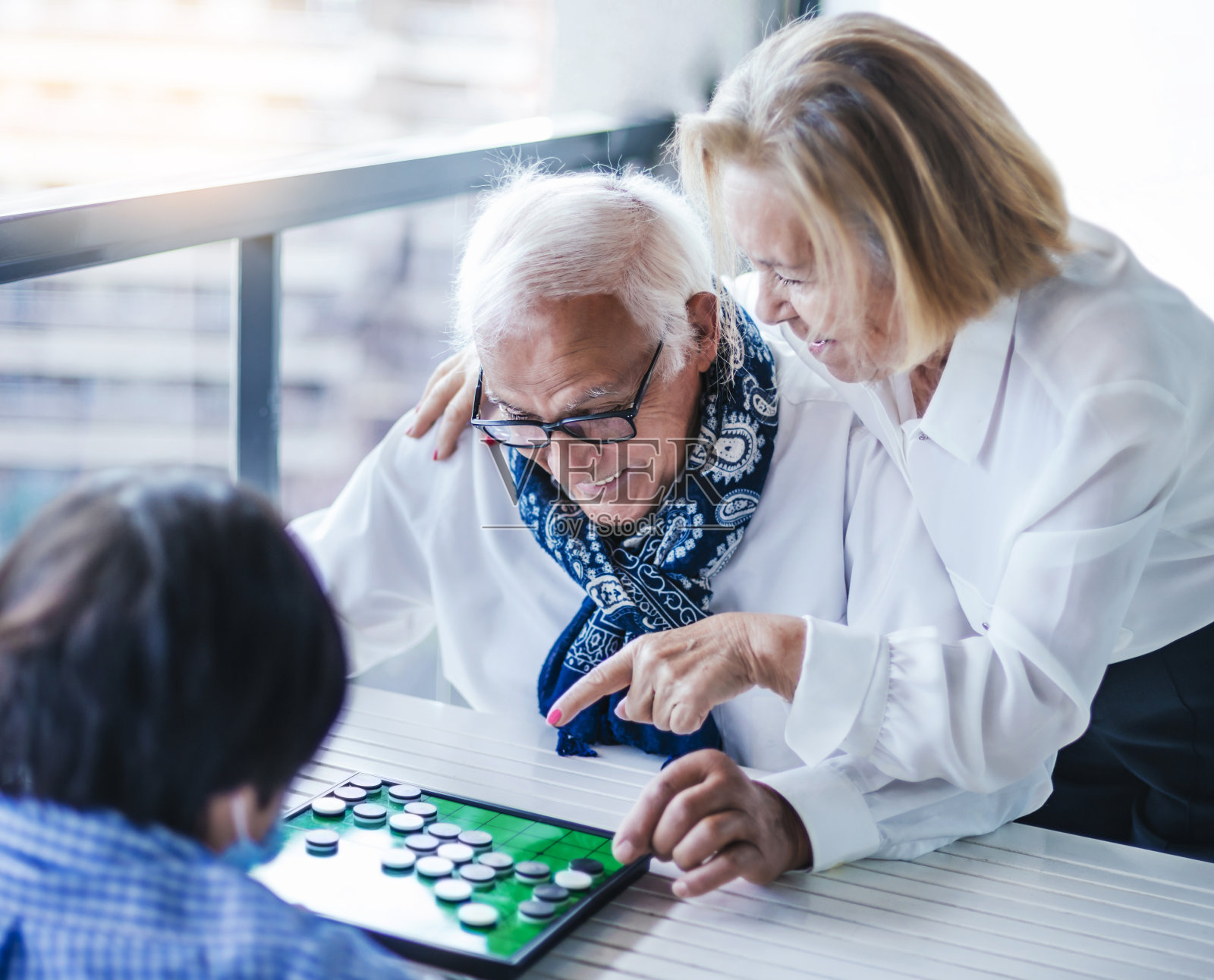 选择性模糊。祖父母和他们的孙子玩棋盘游戏。当他的妻子指出这个游戏时，爷爷笑了。照片摄影图片