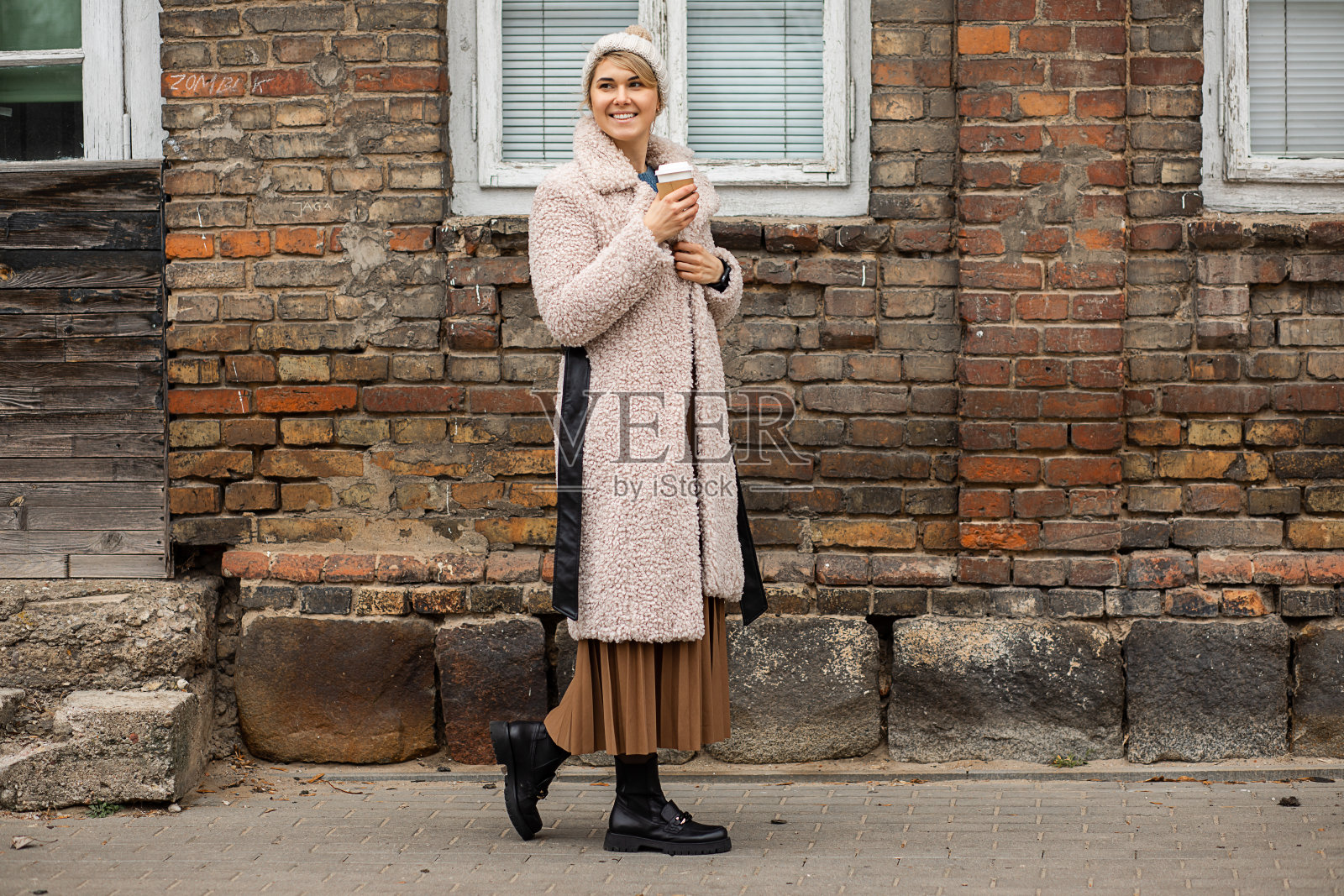 头戴针织帽、身穿人皮大衣的女人，手捧一杯热咖啡，在红色的砖砌旧摊位、街头风格、现代生活的背景下，漫步在城市中照片摄影图片