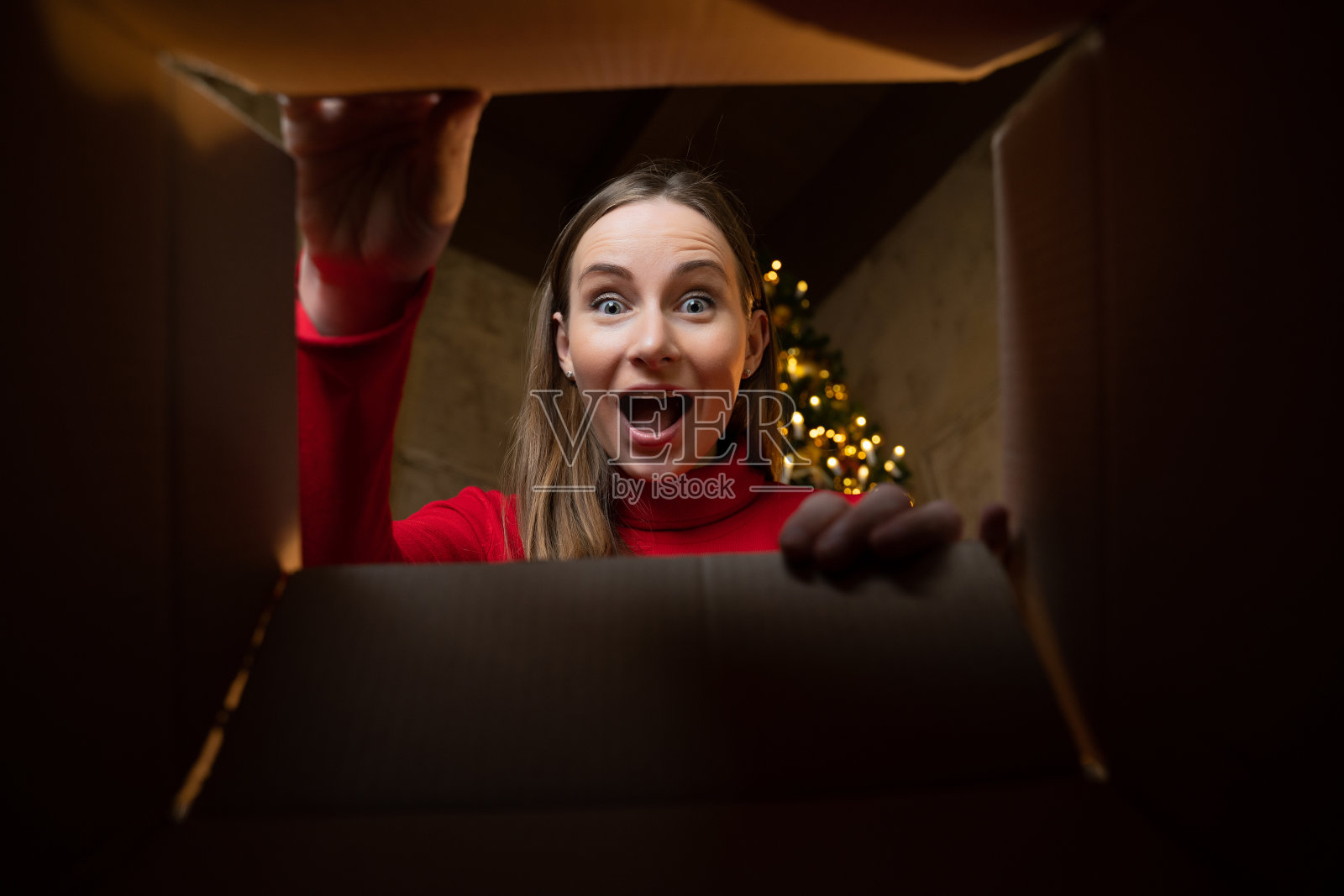 一位身着红色毛衣的欢快漂亮的年轻女子打开了一份圣诞礼物。一个女人在圣诞树旁玩得很开心。从盒子里面看照片摄影图片