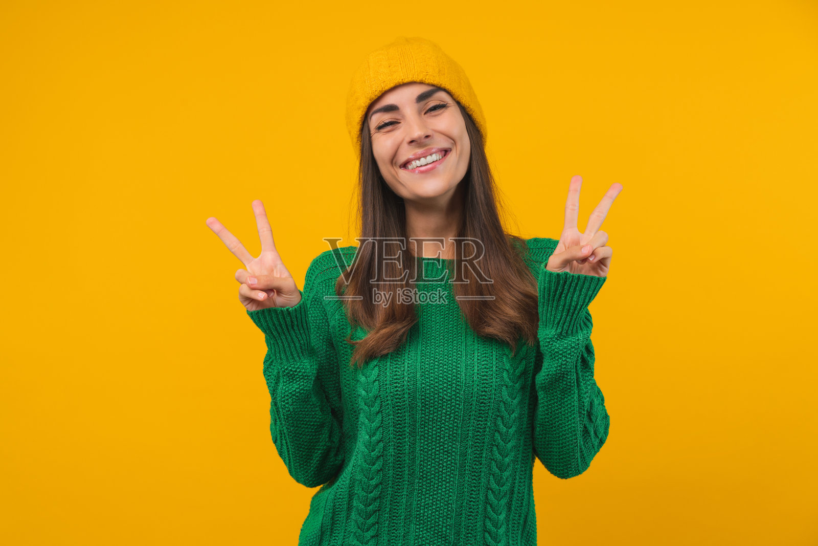 工作室拍摄的年轻快乐的女子穿着绿色毛衣，戴着无檐帽，在明亮的黄色背景下摆出v字形的姿势照片摄影图片