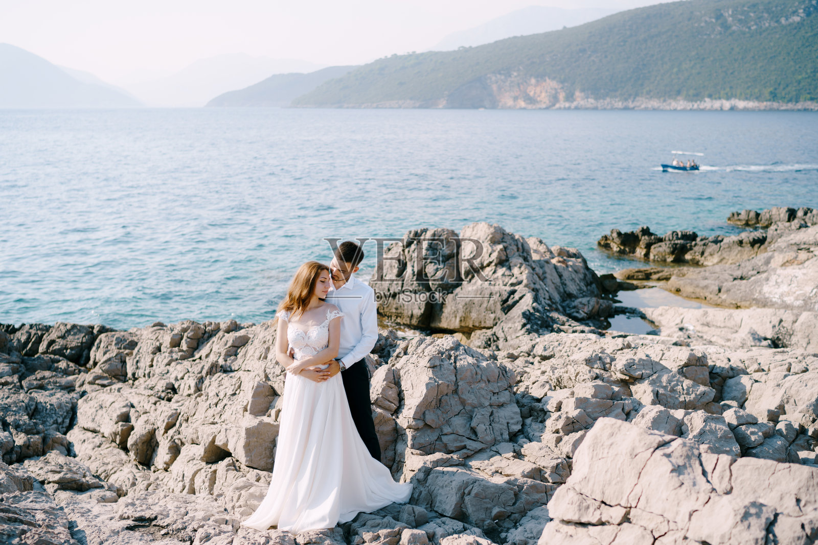 新郎拥抱新娘站在石头上，以大海和山脉为背景照片摄影图片