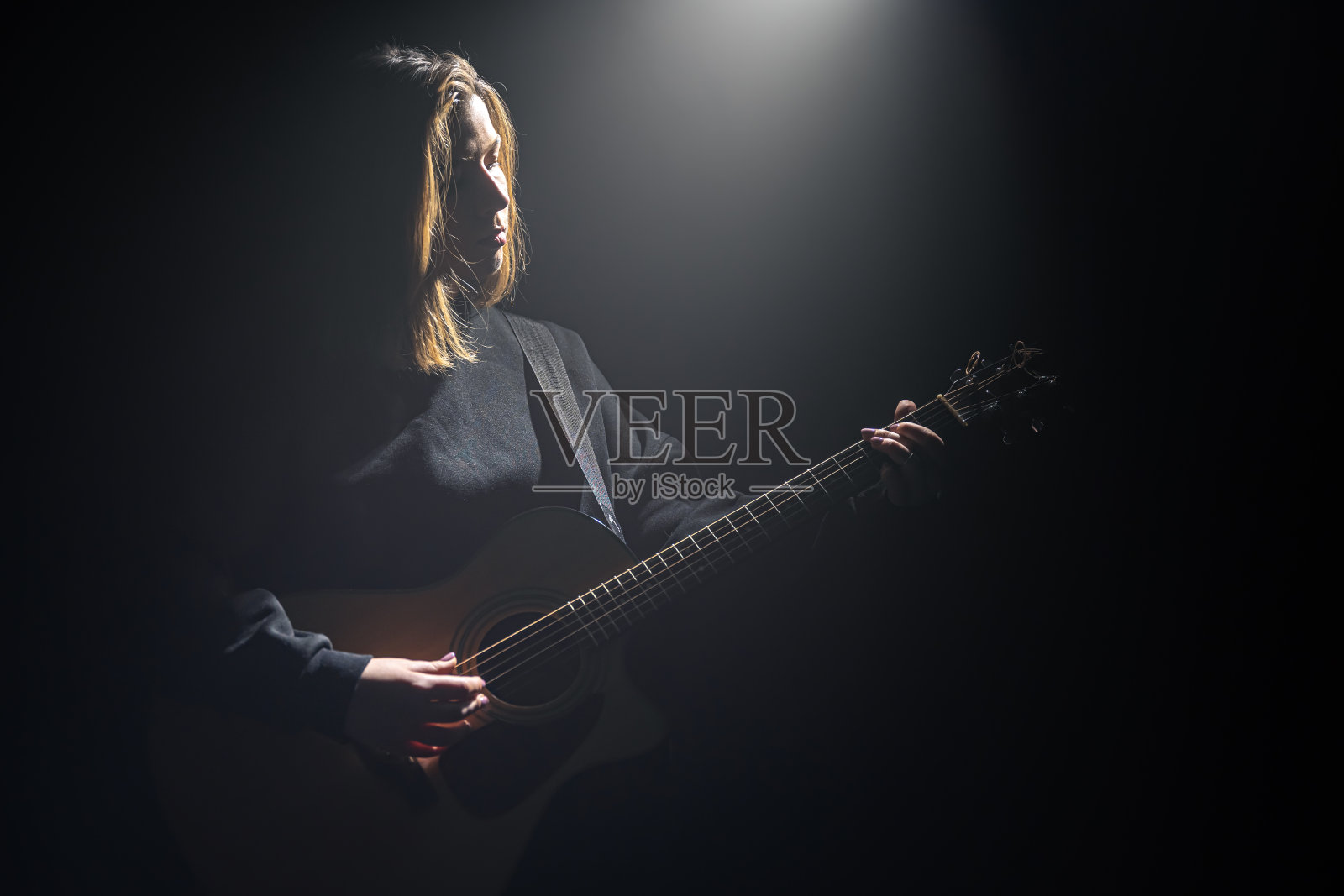 女孩带着一把原声吉他在黑暗中的一束光下。照片摄影图片