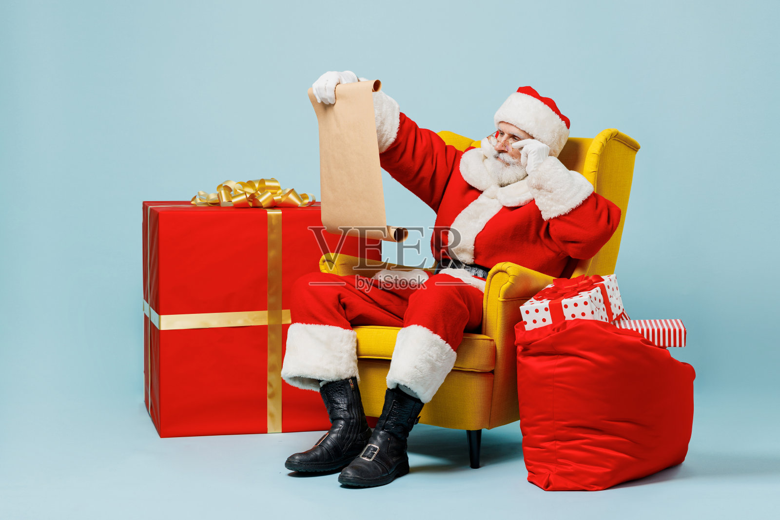 全身上下的圣诞老人50多岁，戴着圣诞帽，穿着红色的衣服，坐在扶手椅上，旁边是礼物袋，读着信，孤立在纯蓝色背景的工作室里。新年快乐，2022年圣诞快乐。照片摄影图片
