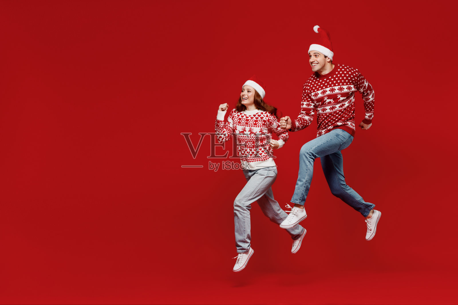 全身年轻的夫妇朋友两个男人女人20多岁在毛衣帽子跳高跑匆忙孤立在纯红色背景工作室肖像。2022年新年快乐庆典，圣诞快乐节日概念。照片摄影图片