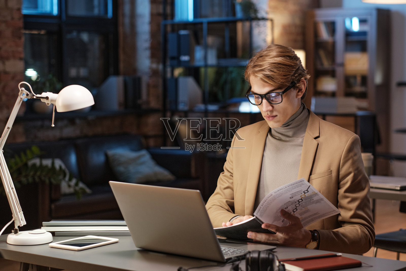 严肃的年轻商人穿着高领毛衣，坐在桌前，拿着笔记本电脑，在黑暗的办公室里分析文件照片摄影图片