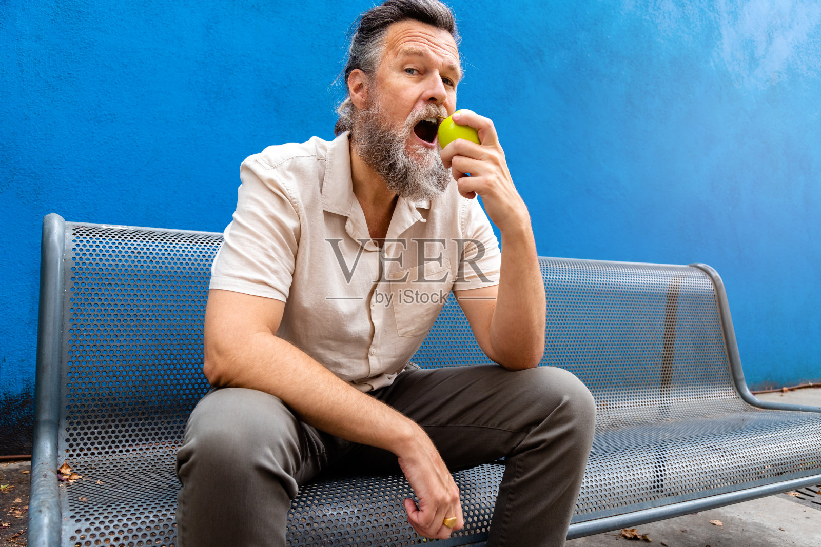 成熟的白种人与胡子吃苹果在户外看着相机。蓝色背景照片摄影图片