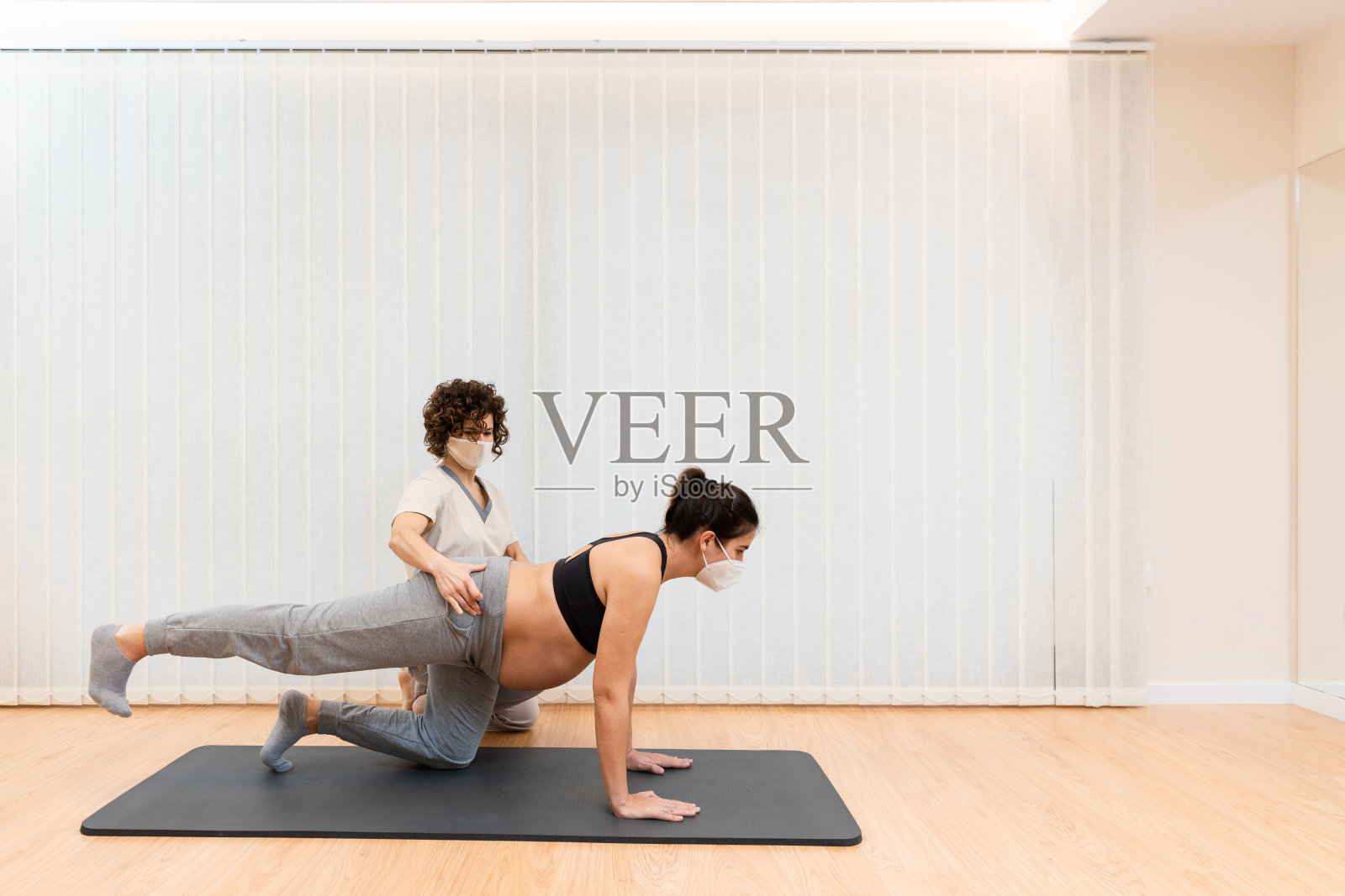 一个带着面罩的孕妇跪在垫子上做普拉提，在理疗师的帮助下有复制空间照片摄影图片