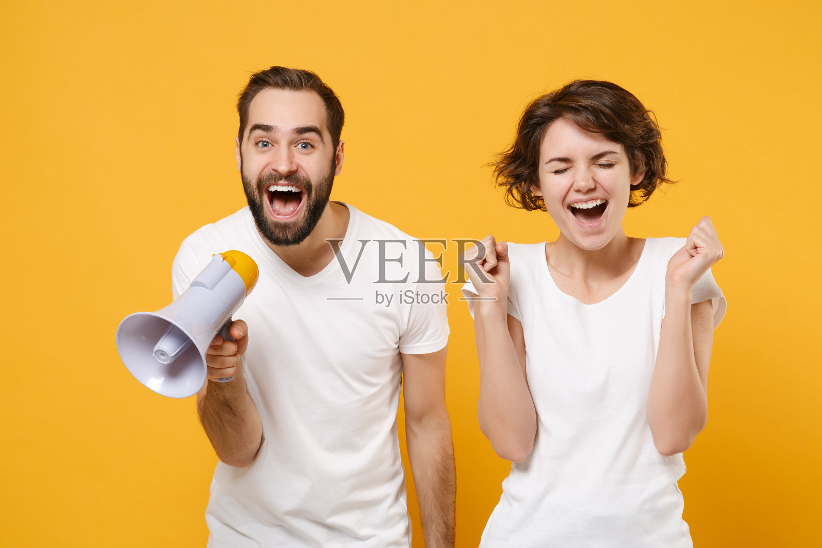快乐的年轻夫妇朋友大胡子的男孩女孩在白色空白的空t恤孤立在黄色橙色的背景。人们生活方式的概念。模拟复制空间。在扩音器里尖叫，做着胜利者的手势。照片摄影图片
