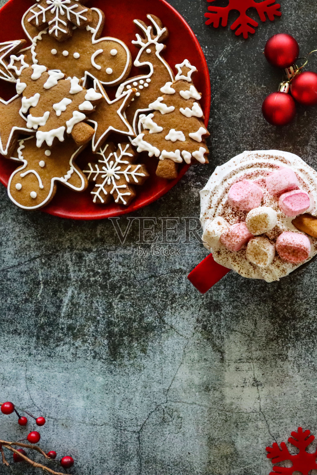近距离图像的红色盘子姜饼人，圣诞树，星星，驯鹿和雪花饼干与白色皇家糖霜，杯子热巧克力，生奶油和迷你棉花糖，红色浆果，小饰品和雪花饰品，灰色背景照片摄影图片