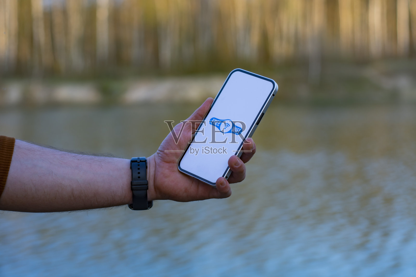 一名男子手里拿着智能手机，屏幕上有一个免费的wi-fi图标。手机背景是森林和湖泊。自然的免费互联网接入。照片摄影图片