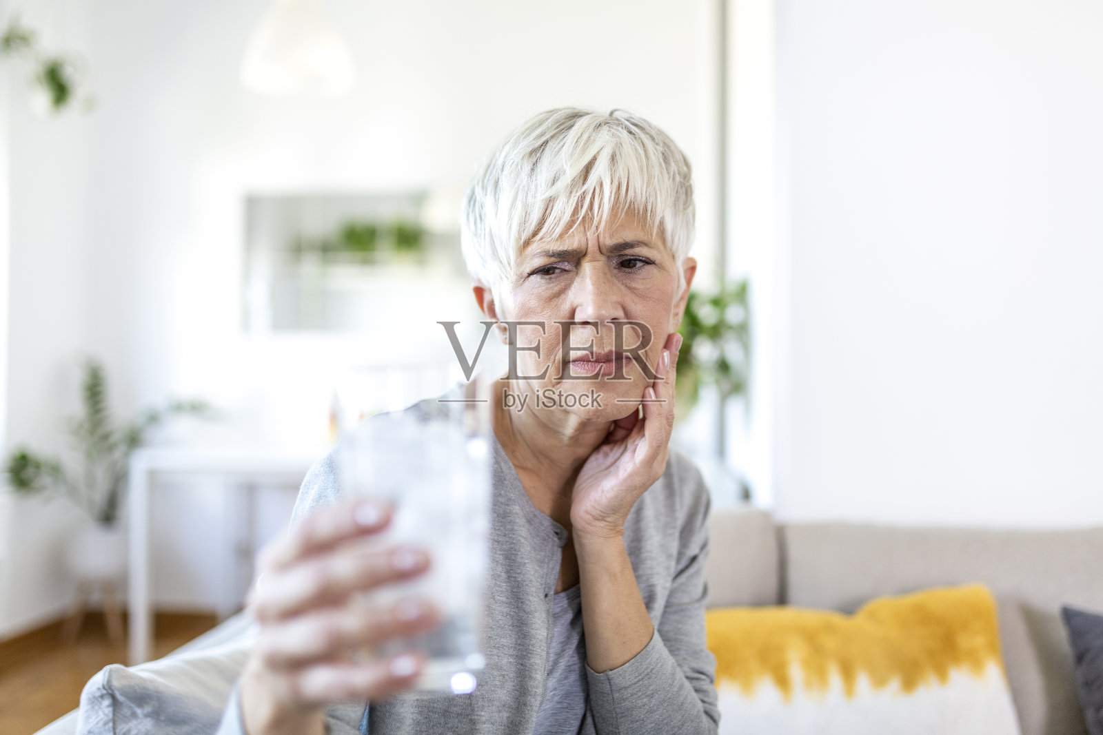 牙齿敏感的老年妇女，手握装有冰的冷水玻璃杯。医疗保健的概念。成熟的女人喝着冷饮，杯子里装满冰块，牙疼，疼照片摄影图片