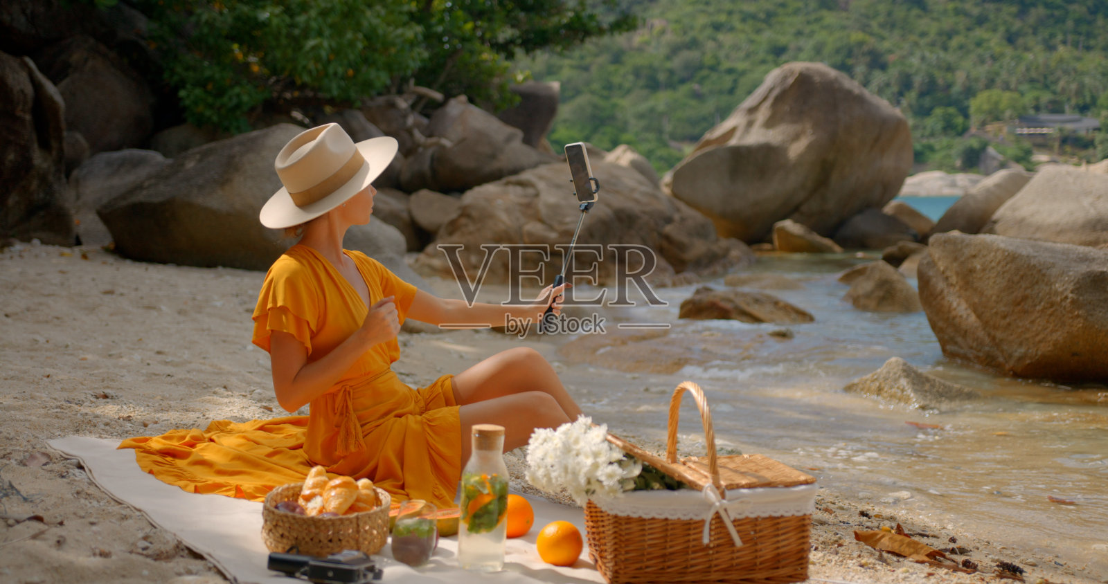 在阳光明媚的日子里，一个穿着黄色衣服的女人坐在孤独的沙滩上，拿着手机，为她的视频博客流媒体照片摄影图片