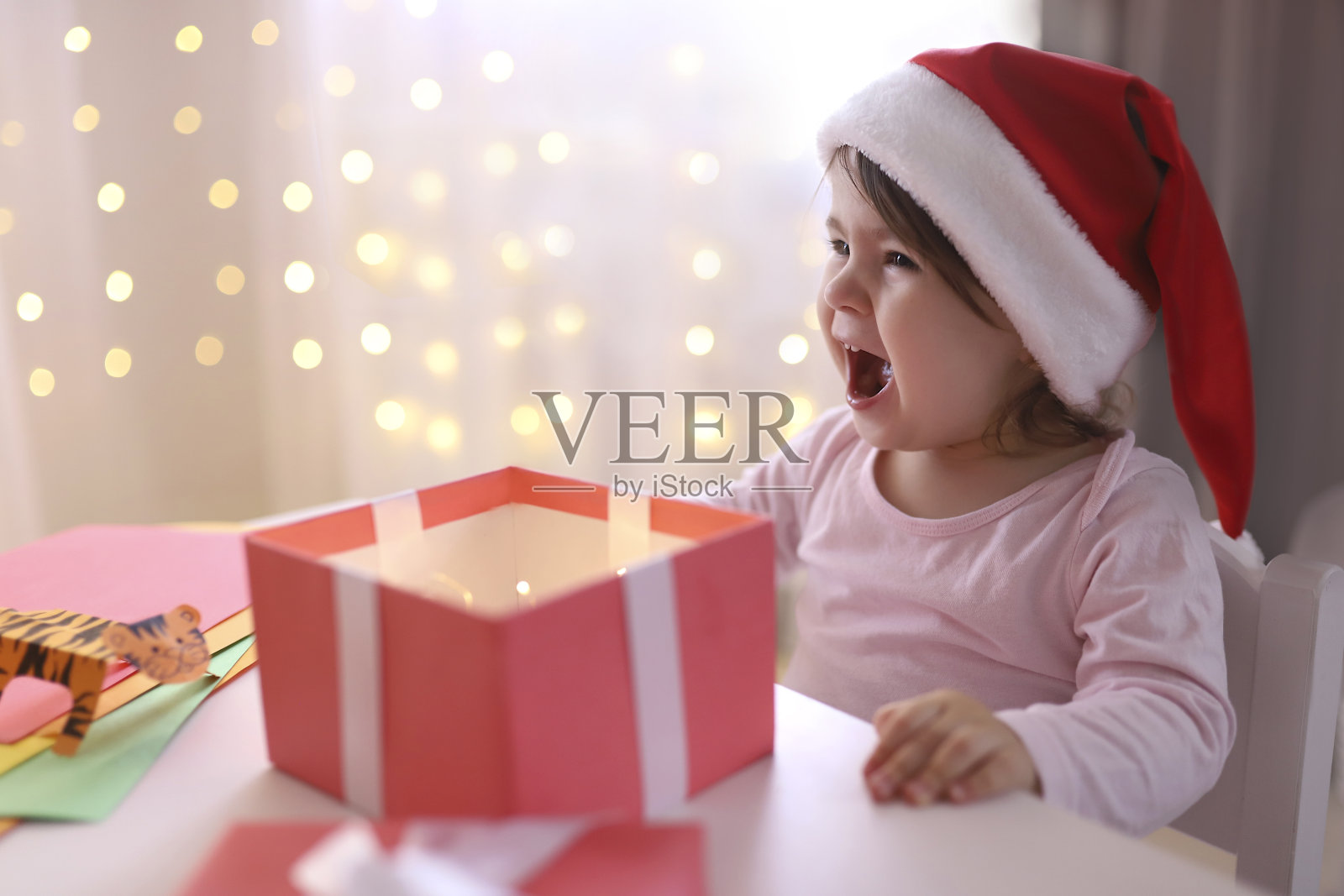 戴着圣诞帽的情绪激动的小圣诞女孩看着礼物盒。孩子得到新年礼物很高兴。背景是散焦的光。2022年圣诞节和新年的概念照片摄影图片