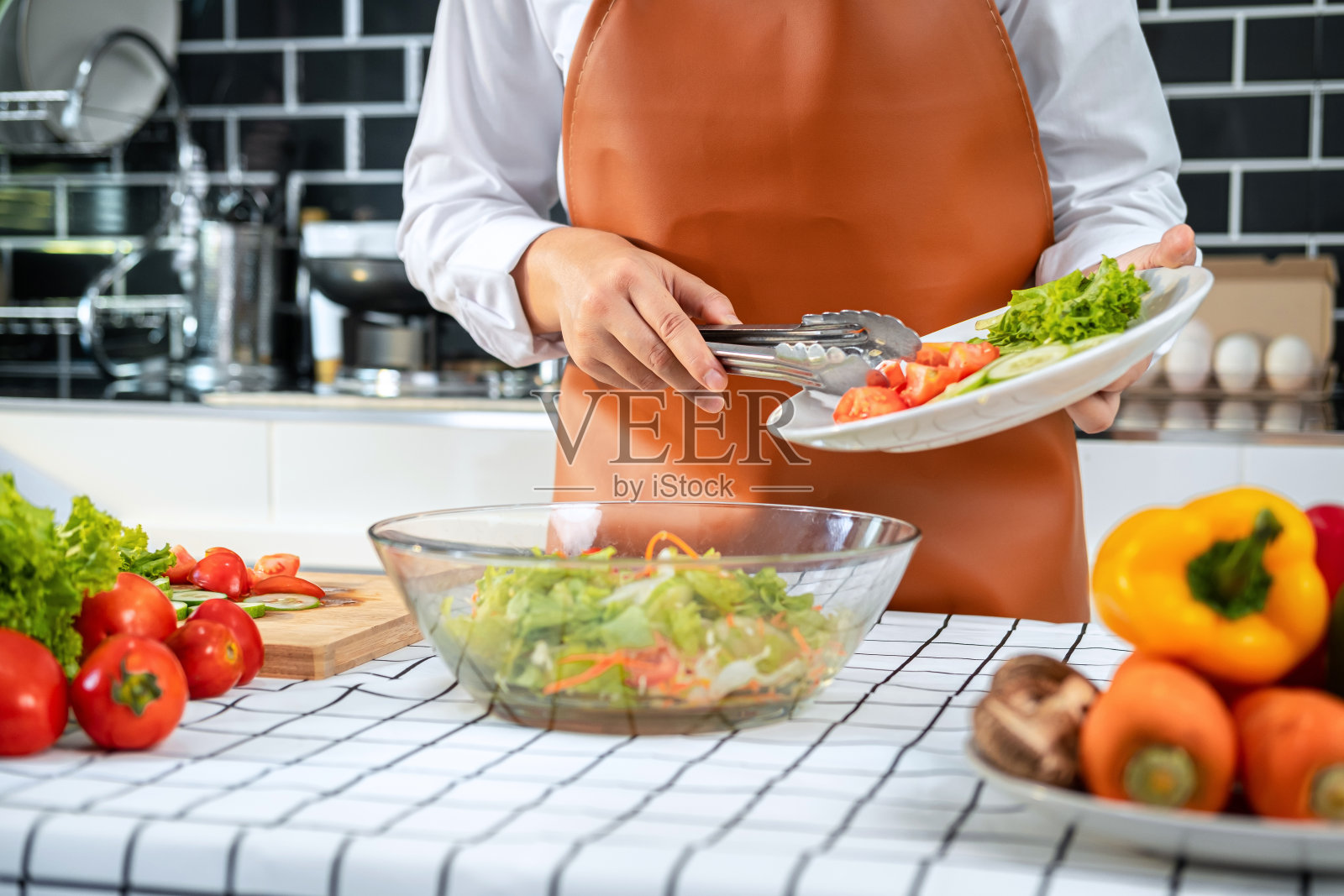 亚洲家庭主妇拿着盛有莴苣、黄瓜和番茄的盘子，用蔬菜钳夹进碗里照片摄影图片