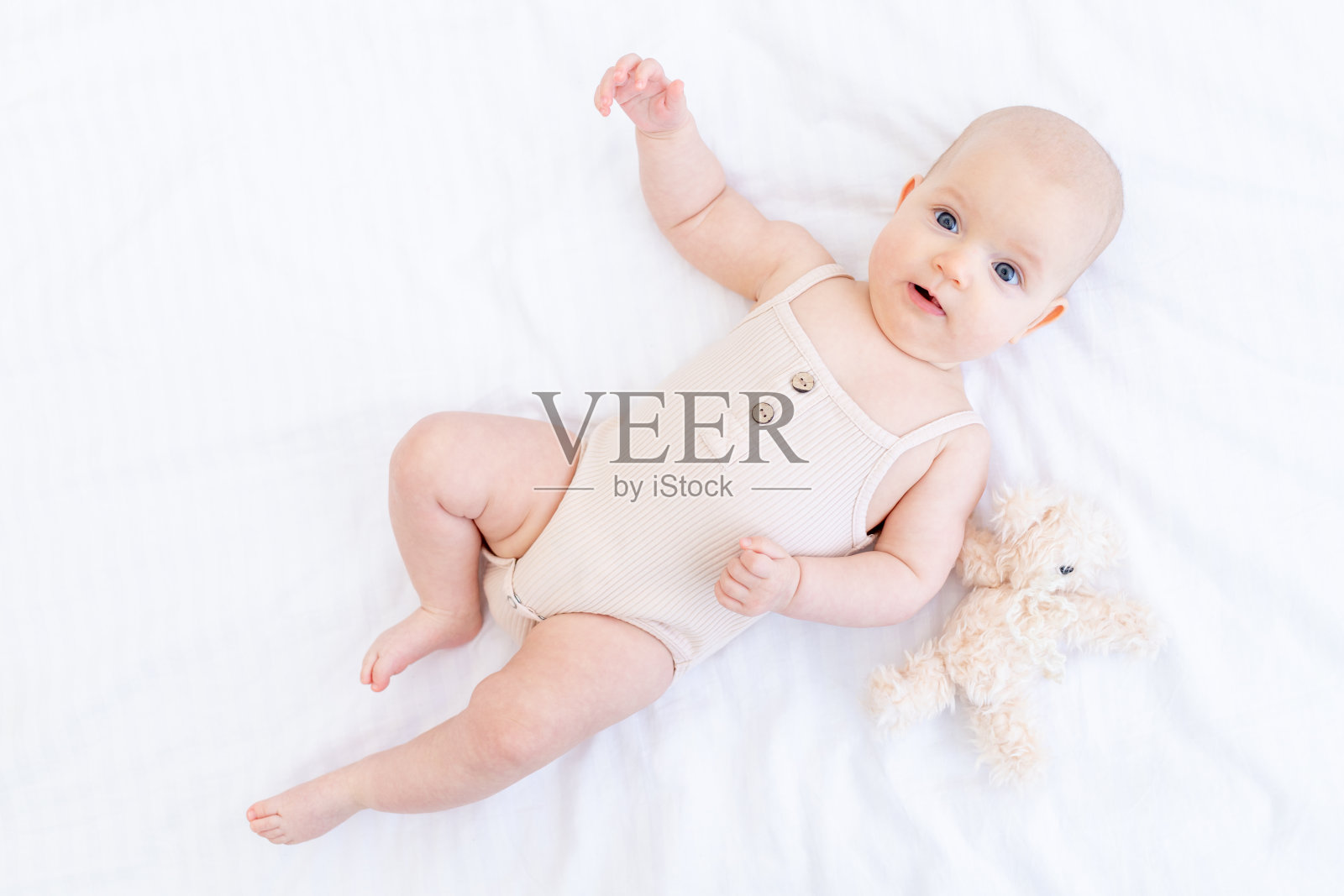 在家中白色的床上，穿着棉质套装的女婴正抱着泰迪熊微笑着躺在床上照片摄影图片