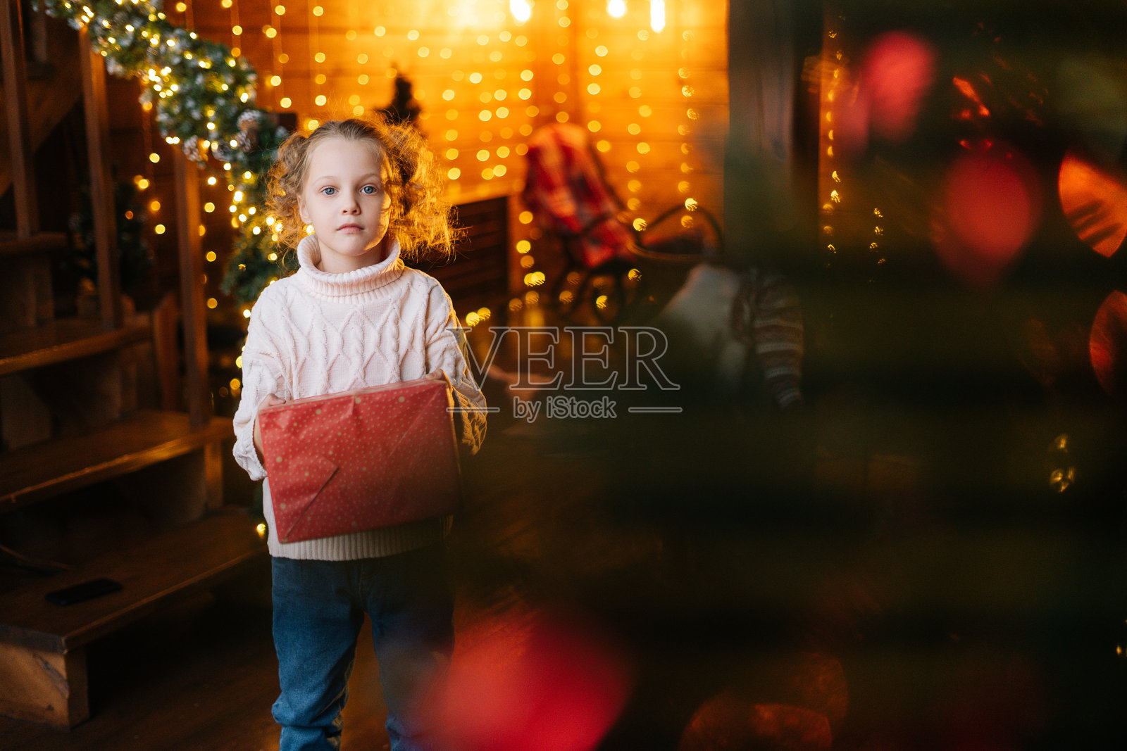 肖像可爱的小金发卷发的孩子拿着红色的礼物盒与圣诞礼物站在舒适的客厅与节日的室内。照片摄影图片