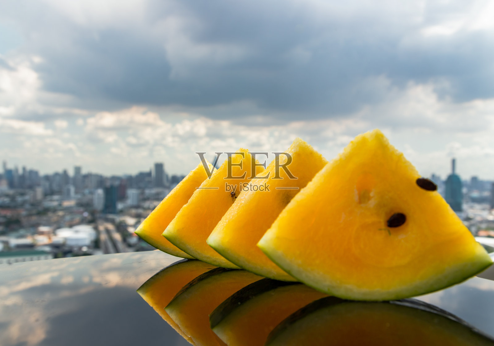 新鲜多汁的黄色西瓜切成三角形在黑色亚克力板反射和城市的看法。照片摄影图片