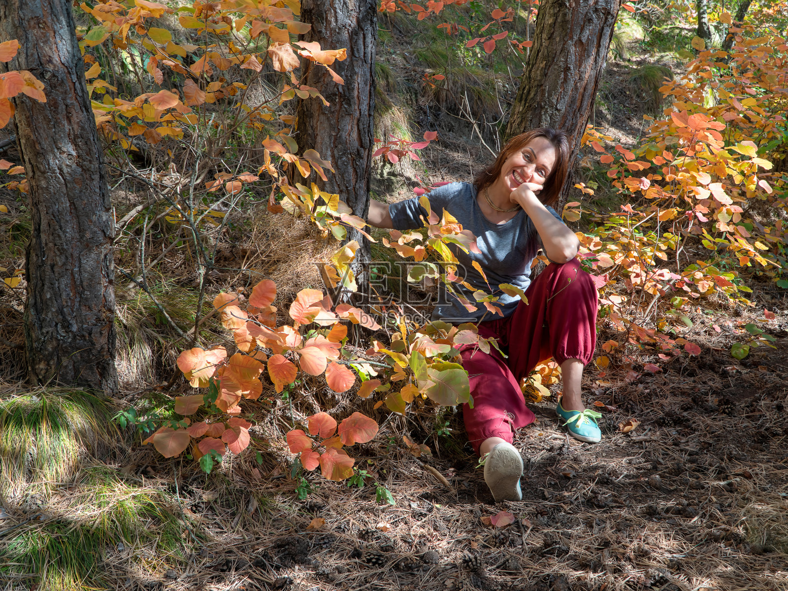 ‌‌穿着红色印度裤的微笑女人坐在森林里，在树叶点缀的小路上的树下照片摄影图片