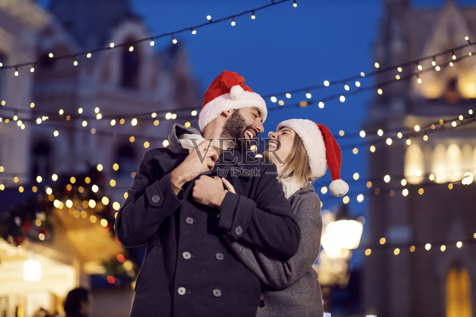 圣诞节前夕，圣诞情侣们在街上笑着拥抱着。一对年轻幸福的夫妇头戴圣诞帽站在街上，在平安夜拥抱在一起。照片摄影图片