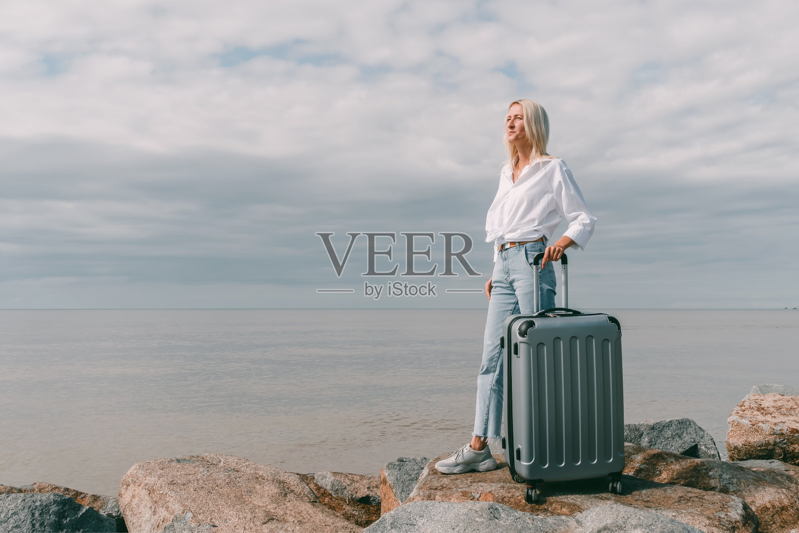 一个年轻的金发女人，穿着牛仔裤和白衬衫，提着一个手提箱，在阴天里站在海边的巨大岩石上，看着别处照片摄影图片