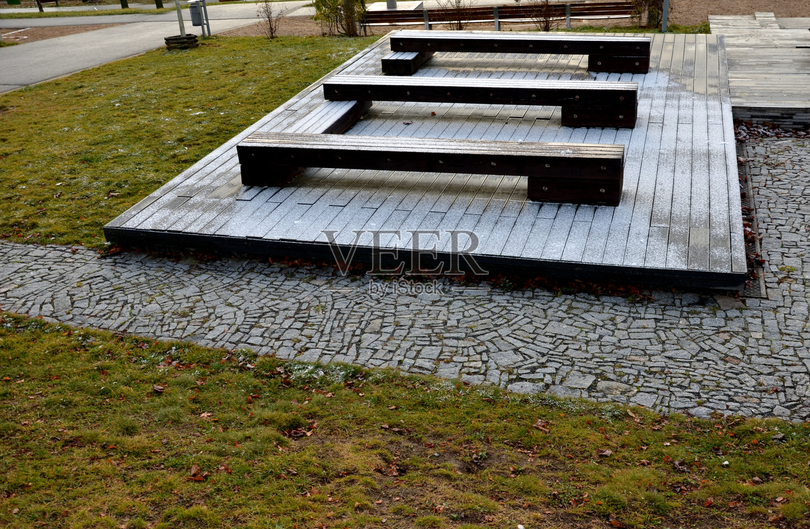 公园里的木制露台和棕色的螺丝固定的长凳把zig一个接一个地放回去。这些长椅是一个挨着一个的。固定在金属格栅上的树照片摄影图片