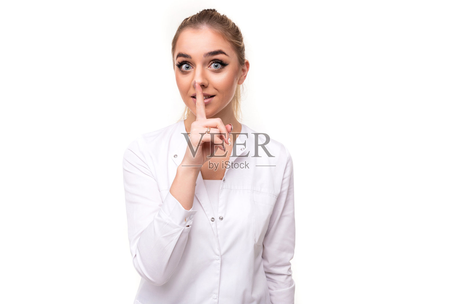照片是一个漂亮的漂亮的可爱的有吸引力的积极的年轻金发女人，马尾辫和化妆在一个护士的白色医疗大衣嘘和秘密的手势，一个手指孤立在白色的背景上，自由和复制空间的文本照片摄影图片
