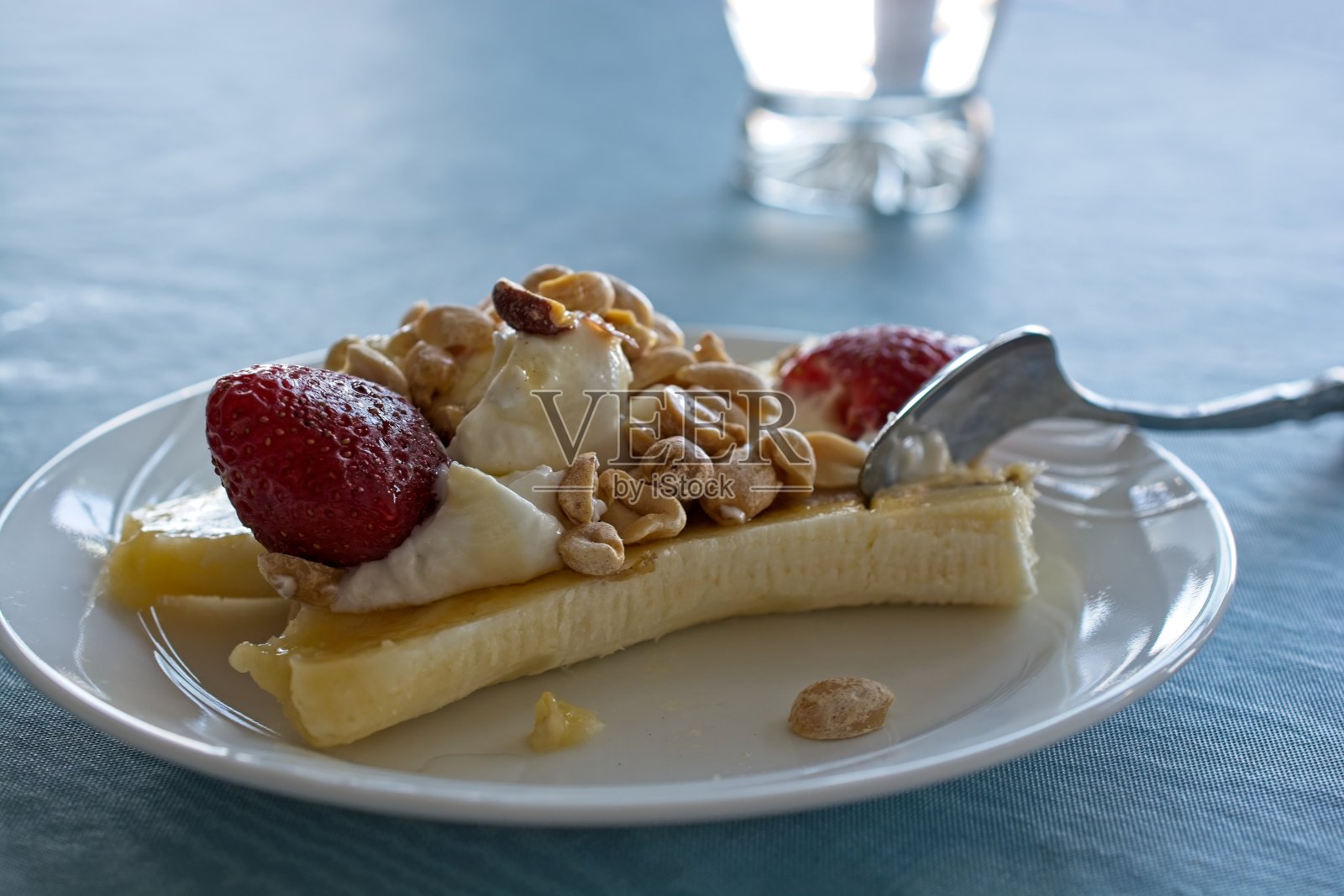 香蕉船放在蓝色桌布上的白色盘子里，背景是勺子和一杯水照片摄影图片