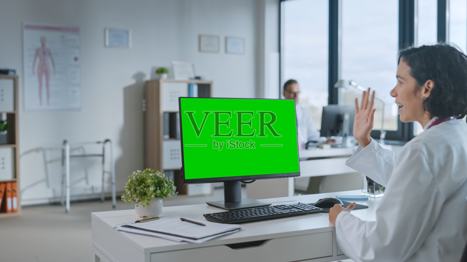 一名女医生正在健康诊所的一台绿屏模拟显示器上与病人进行视频通话。穿白大褂的助理在医院办公室谈论健康问题。照片摄影图片