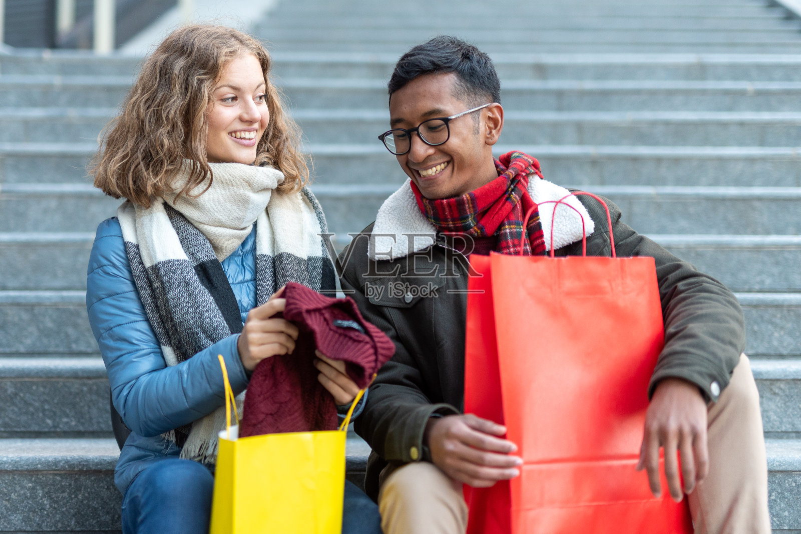 多种族订婚的年轻夫妇坐在城市的楼梯上看着他们的购物，金发女人在她的北非裔男友旁边展示一件新衣服照片摄影图片