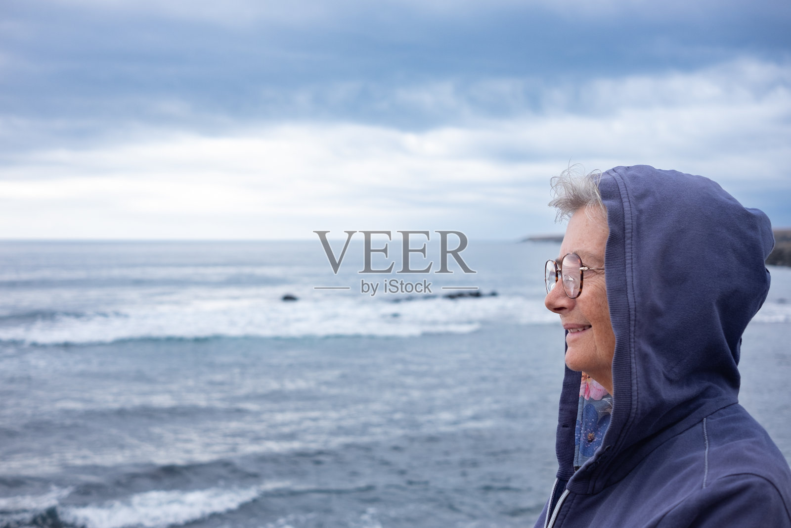 一个头上戴着兜帽的年长女人在阴天里站在海边，望着远方。70多岁的老年妇女享受退休和自由照片摄影图片